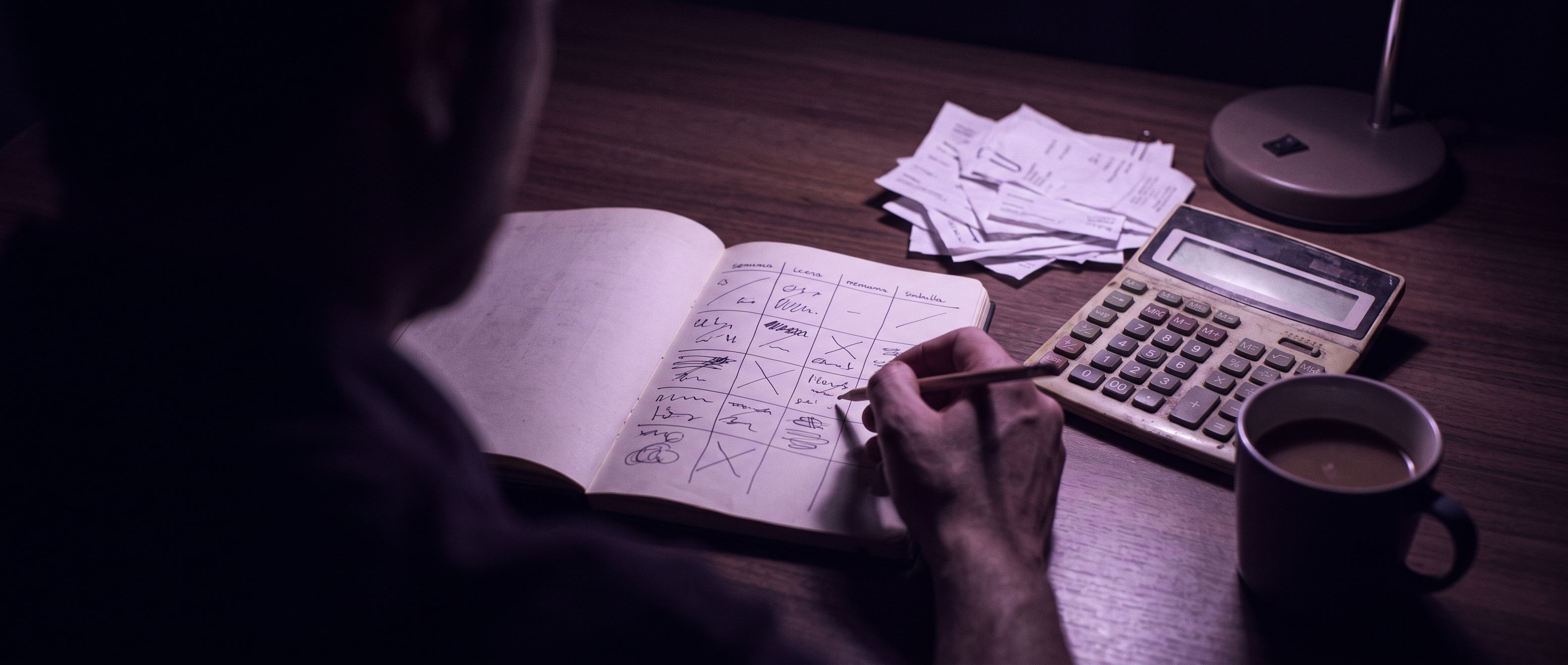 Una persona revisa de noche una libreta con una tabla por semanas, junto a una calculadora, facturas y una taza de café.