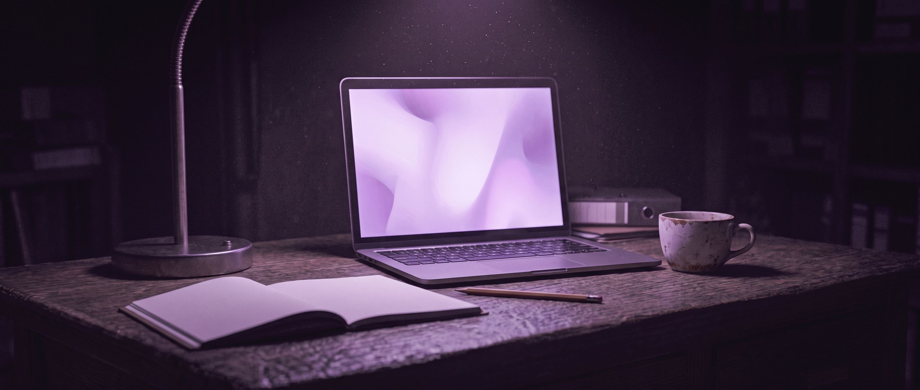Un portátil abierto en un escritorio de noche, con la pantalla iluminando una libreta, un lápiz y una taza.